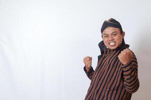 Portrait of excited Asian man in javanese cloth with blangkon hat raising his fist, celebrating success after winning a competition. Isolated image on white background photo