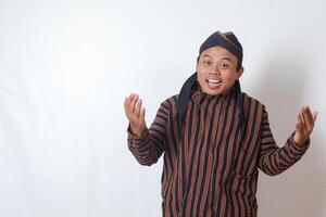 Portrait of excited Asian man in javanese cloth with blangkon hat pulling hands towards camera, inviting someone to come inside, welcoming gesture. Isolated image on white background photo