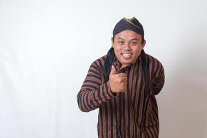 Portrait of amazed Asian man in javanese cloth with blangkon hat showing approval and agreement sign, making thumbs up hand gesture. Isolated image on white background photo