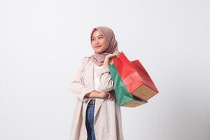 Portrait of cheerful Asian hijab woman in casual suit holding and showing colorful paper shopping bags. Shopaholic girl and discount buying concept. Isolated image on white background photo