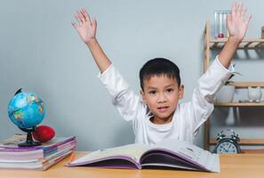 Concept of education and homework and school. Little boy smiling and raising his hand happily while sitting at a table with a book. Children celebrating finishing their homework. photo