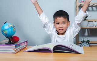 Concept of education and homework and school. Little boy smiling and raising his hand happily while sitting at a table with a book. Children celebrating finishing their homework. photo