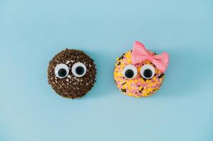 Two sweet cookies covered with colorful sprinkles with googly eyes and one with pink hair bow on light pastel blue background. Minimal sweet food concept. Trendy funny biscuits idea. Flat lay. photo