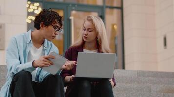 Young asian guy with a copybook and pen asking questions about work to a young caucasian woman with a laptop. Colleagues sitting on stairs near a building and discussing working issues video