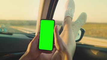 A girl in the front seat of a car uses a phone with a green screen, her feet out the window. Top view of her hands holding the phone with a chromakey. video