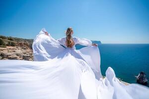 Blonde with long hair on a sunny seashore in a white flowing dress, rear view, silk fabric waving in the wind. Against the backdrop of the blue sky and mountains on the seashore. photo