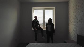 Silhouetted couple sitting by a window in a minimalistic room, A couple sits on the windowsill of a minimalistic, sparsely furnished room, silhouetted against the light coming through the window video