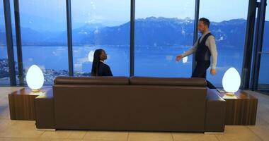A person is seated on a sofa, looking at mountains and a lake through windows in a modern room video