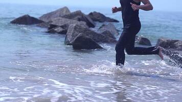 A man is seen running along the shoreline of a rocky beach, dressed in a black wetsuit video