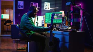 Musical producer prepares to record tunes on acoustic instrument, working in personal home studio to record soundtrack. Sound composer recording guitar music, editing with mixing console. Camera B. video