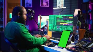 Young musician mixing and mastering songs next to isolated mockup display, operating on digital audio software to add sound effects on new tracks recorded. Audio engineer in home studio. Camera B. video