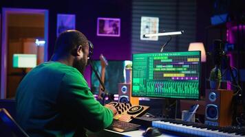 African american composer creating a new track for a project, using professional production pc interface in his home studio. Creative artist songwriter producing music with sound effects. Camera B. video
