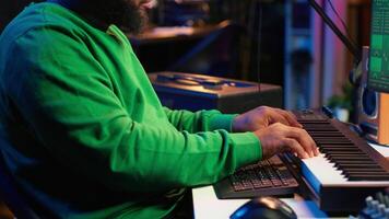 Songwriter mixing and mastering tracks on stereo console, playing electronic keyboard in home studio. Artist oversees audio performances to create melody, producing tunes with piano keys. Camera A. video