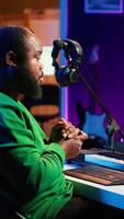 Vertical African american sound engineer using midi controller notes to add audio effects in post production, editing tracks with digital software and mixing console. Male musician in home studio. Camera A. video
