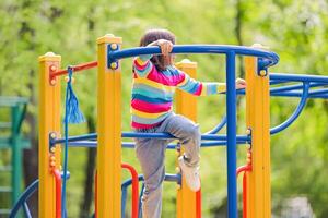 A little girl 5-6 years old, in a bright striped jumper, climbs on a multi-colored playground, in a park photo