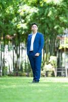 Asian handsome glasses man in a blue suit and the same color pants is looking at the camera, smart smiling and standing in the garden. photo