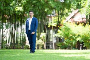 Asian handsome glasses man in a blue suit and the same color pants is looking at the camera, smart smiling and standing in the garden. photo