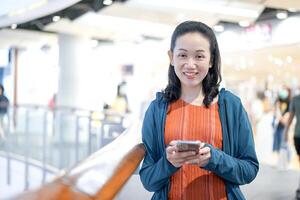 Smiling Asian Female Looks at the camera with holds smartphone in hand, Stands in Department Store Atmosphere. photo