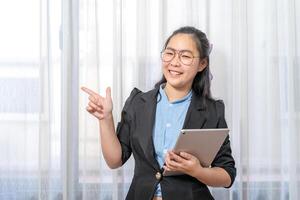 Happy Asian woman fingering up and pointing with tablet computer on her hand at copy space while standing in front of the transparent curtain see through the window light. photo
