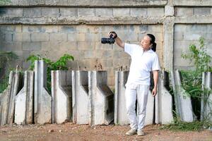 Professional Asian Chinese - Thai Camera man posture and focus in viewfinder and LED screen on mirrorless camera medium format in construction concrete area. photo