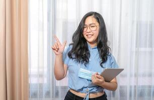 Happy Asian woman fingering up and pointing with tablet computer on her hand at copy space while standing in front of the transparent curtain see through the window light. photo