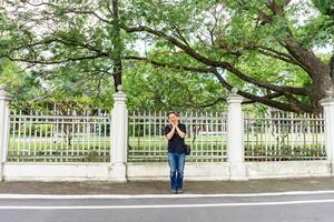 Asian Black T-Shirt is standing in front of white vintage fence beside the road in the afternoon time. photo