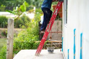 Man who wearing dark pants is climbing a red stairs to fix the house. Green shrub and broken pillar are on the background photo