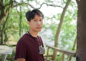 Asian Smart man in casual dark red T-shirt and hang on the gray cloth mask on his neck, stands and looks to the camera with the green tree blur background. photo