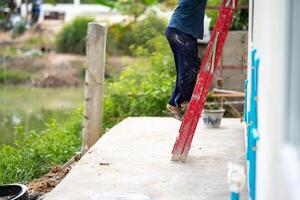 Man who wearing dark pants is climbing a red stairs to fix the house. Green shrub and broken pillar are on the background photo