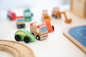 A Toy of wooden school bus drives beside a green car with many kinds of car behind it of a Vehicle Toy Set for children in an indoor playground. photo