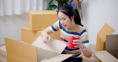 Handheld high angle view shot, Young Asian woman unpacking cardboard boxes and packing things out of the boxes to prepare for decorating in new house. Moving house and lifestyle concept. video