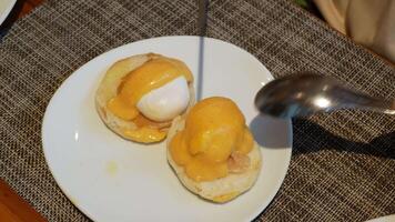 using knife and fork cutting egg yolk to eat egg benedict onn english muffin with beacon in white plate. video