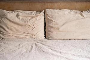 Two beige pillows on an unmade bed with wrinkled sheet and headboard. photo
