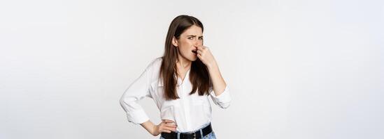 Disgusted adult woman, 25 years old, shut nose from dislike and disgust, bad smell, standing over white background photo