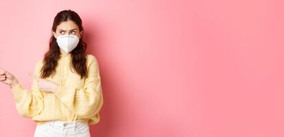 Covid, corona and social distancing concept. Confused brunette girl in medical respirator having doubts, pointing and looking aside at left copy space, standing against pink background photo