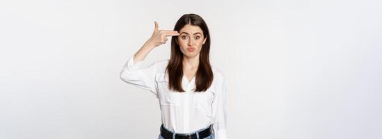 Image of annoyed young woman showing finger gun gesture over head, bothered by smth, tired, standing over white background photo