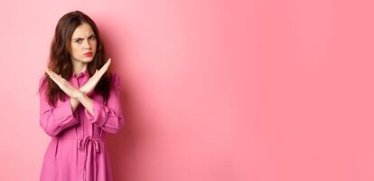 Displeased angry woman blocking offer, showing cross stop gesture, saying no and shaking head in negative reply, standing over pink background photo