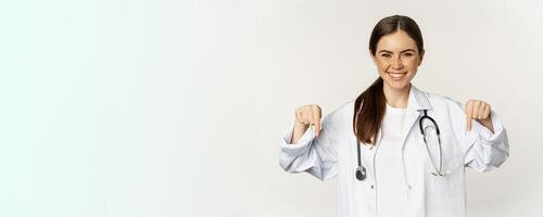 Portrait of happy female doctor, pointing fingers down and smiling, demonstrating promo offer, discount, standing over white background photo