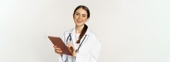 Doctor in white coat using digital tablet, reading medical data on gadget, working in hospital, standing over white background photo