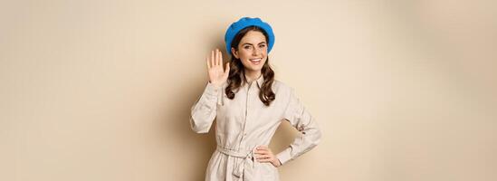 Image of beautiful friendly woman in outerwear, saying hello, waving hand and smiling, greeting you, standing over beige background photo