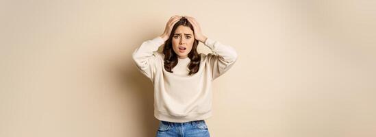Frustrated and troubled woman facing disaster, holding hands on head in panic, looking anxious, standing over beige background photo