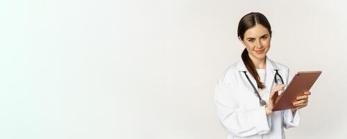 Doctor in white coat using digital tablet, reading medical data on gadget, working in hospital, standing over white background photo