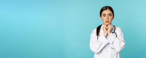 Worried and concerned physician, hospital worker in white lab coat, looking scared and nervous at camera, standing with stethoscope over torquoise background photo