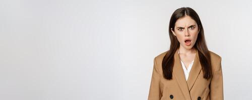 Frustrated, insulated woman in business outfit, staring startled and shocked at camera, standing over white background photo