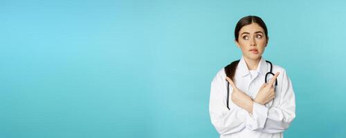 Portrait of indecisive, confused female doctor, pharmacy worker pointing sideways and biting lip clueless, dont know, standing over blue background photo