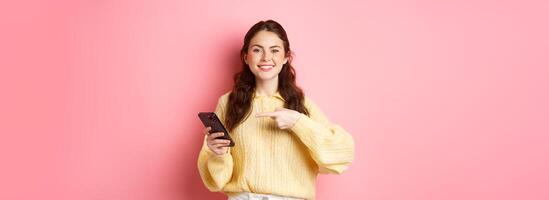 imagen de feliz, sonriente joven mujer, participación móvil teléfono y señalando a móvil pantalla con satisfecho y confidente rostro, en pie terminado rosado antecedentes foto