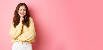 Portrait of smiling beautiful lady, looking determined, touching chin and staring satisfied, seeing something interesting, have idea, standing thoughtful against pink background photo
