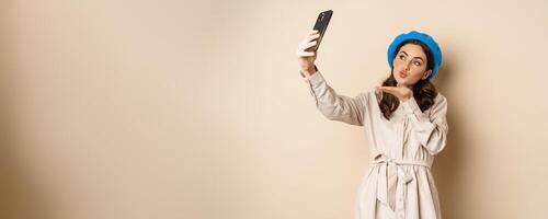 hermosa moderno mujer posando para autofoto, participación teléfono inteligente, tomando imagen foto en móvil teléfono, en pie contento en contra beige antecedentes