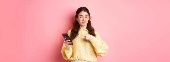 Skeptical young woman grimacing unsatisfied, pointing finger at smartphone screen, having doubts about online content, standing against pink background photo