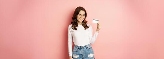 Portrait of attractive smiling woman showing credit card, recommending app, store with discounts, standing against pink background photo